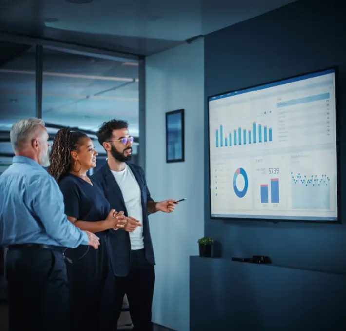 Three people in business attire stand in an office, looking at charts and graphs displayed on a large screen during a presentation.