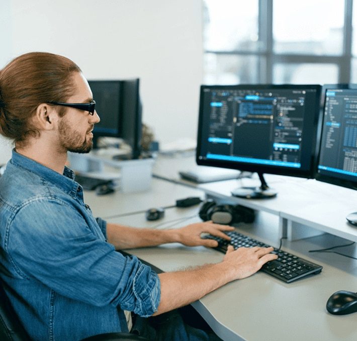 A person with a ponytail and glasses works at a desk with two monitors displaying code in a modern office environment.