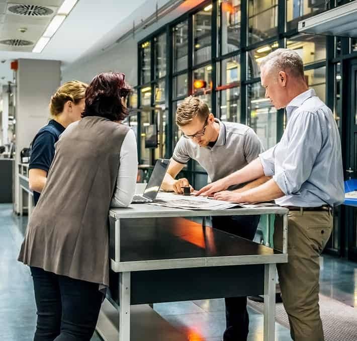 Four people stand around a table in a modern office or laboratory, examining documents and a laptop, engaged in discussion.