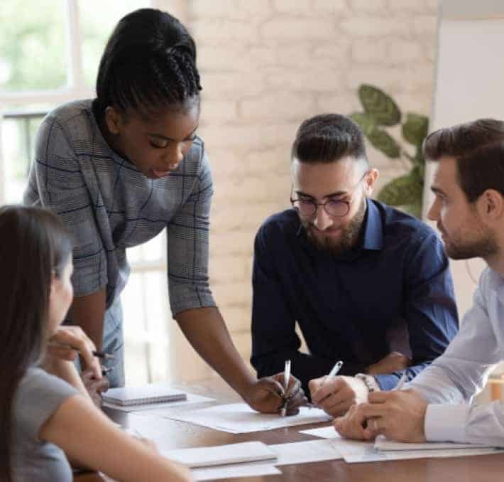 Four people sit around a table discussing documents; one woman stands and points at a paper while the others listen and take notes.