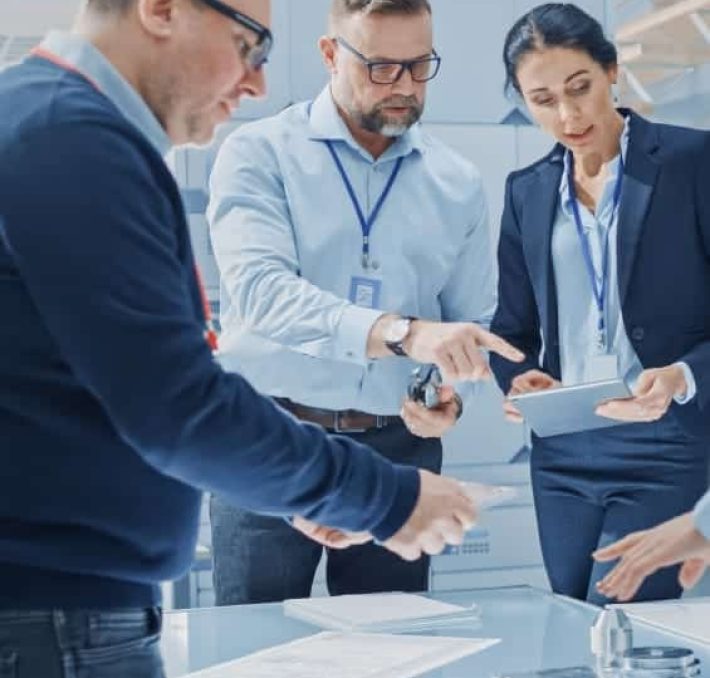 Three professionals in business attire stand around a table, reviewing documents and a tablet in an office setting.