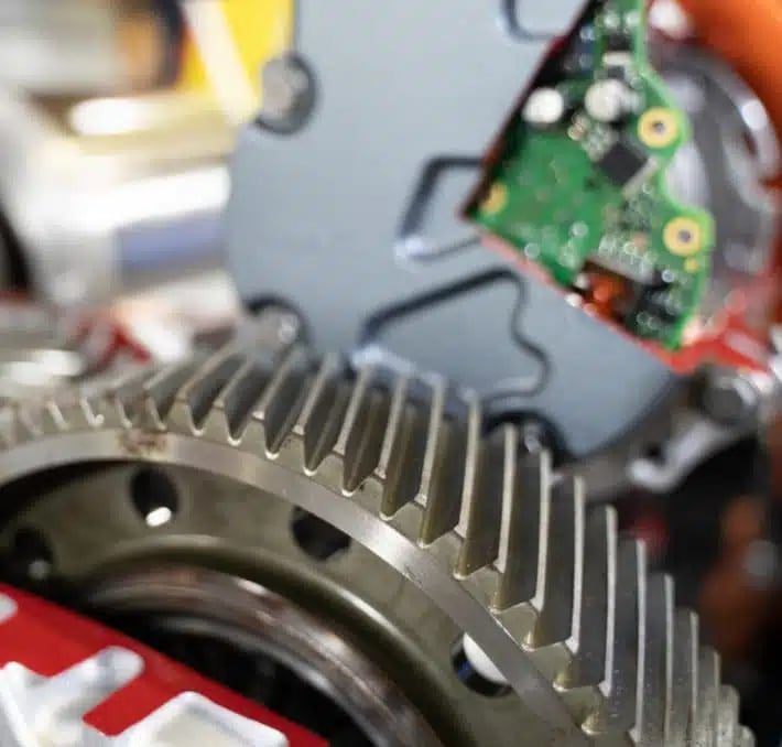 Close-up view of a large metal gear with a circuit board and mechanical components visible in the background.