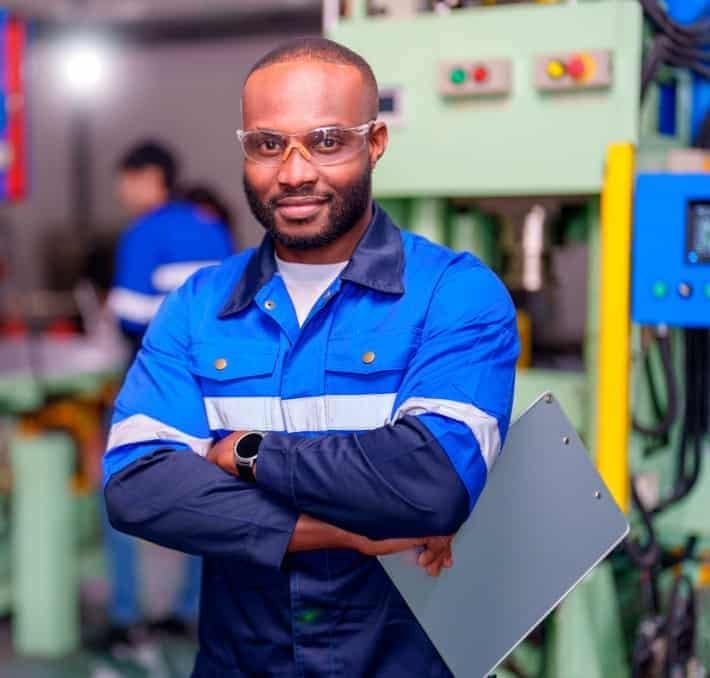 Man in blue work uniform and safety glasses stands with crossed arms, holding a clipboard, in an industrial or factory setting.
