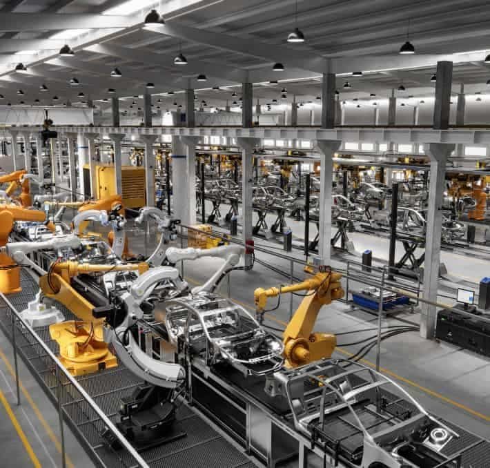 Automated robotic arms assemble car bodies on a production line inside a large, modern automotive factory.
